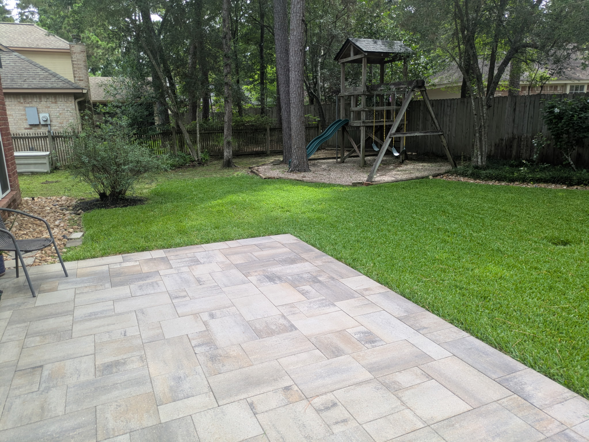 A back garden in Montgomery County with a mown lawn running up to a paver patio and a wooden play set under the pines